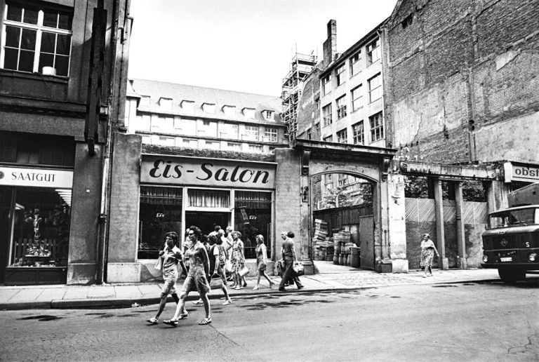 An einer Straße mit Kriegsschäden an den Gebäuden sind vor einem Eis-Salon ein paar junge Menschen unterwegs.