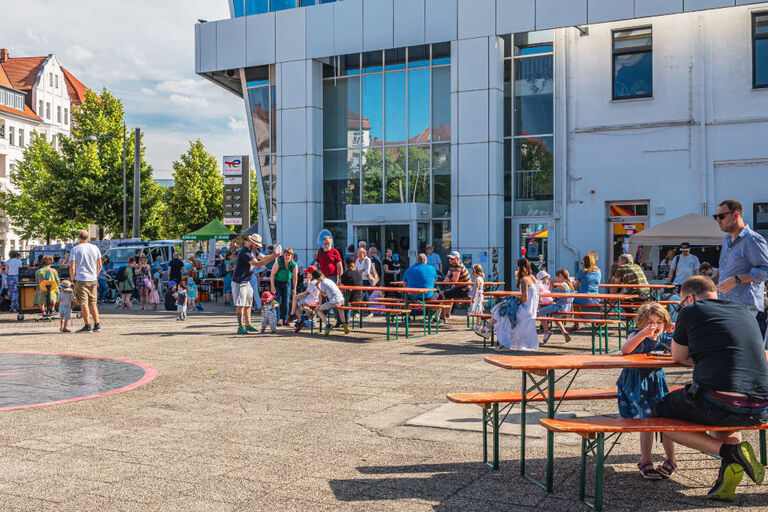 Vor einem großen Glasgebäude sind viele Menschen zwischen Bierbänken und Ständen unterwegs.