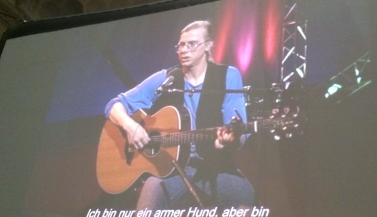 Gerhard Gundermann sieht man auf einer Leinwand Musik machen