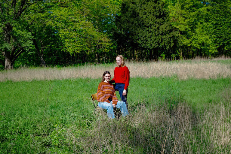 Zwei junge Frauen auf einer großen Wiese, wobei eine von ihnen auf einem Stul sitzt