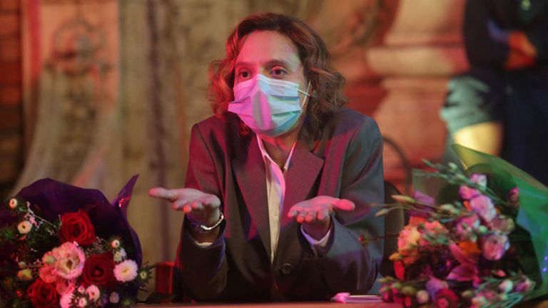 Szene mit der Haupdartellerin, die mit medizinischer Maske zwischen zwei Blumensträußen an einem Tisch sitzt.