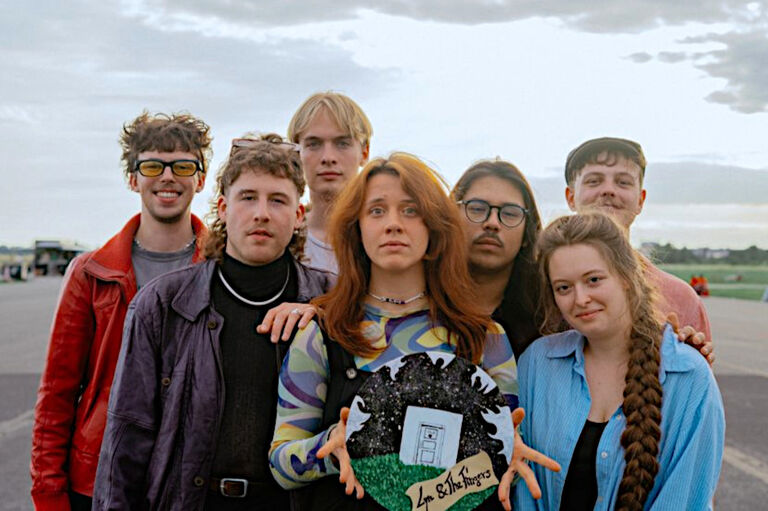 Gruppenfoto der ernst blickenden Band auf einem weiten betonierten Platz unter einem wolkenverhangenen Himmel