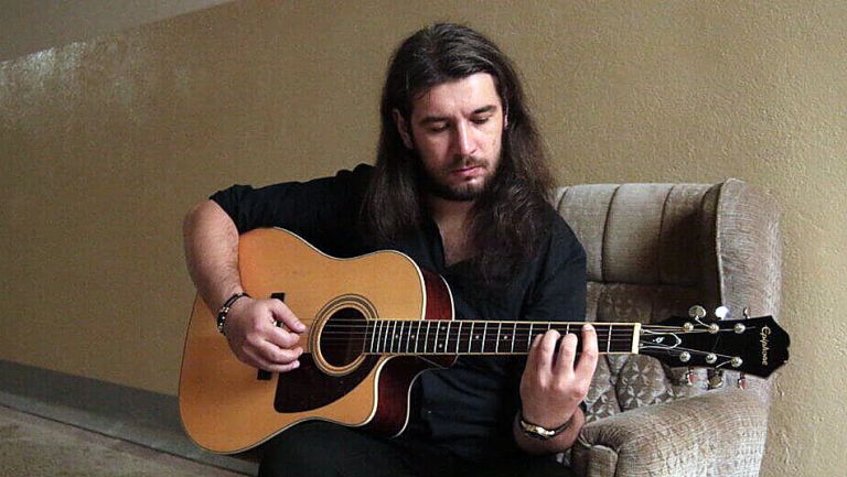 Der Musiker sitzt in einem großen kahlen Raum auf einem alten Sessel und spielt Gitarre.