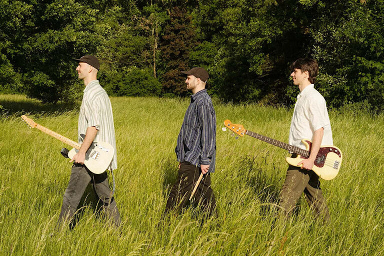 Die drei Musiker laufen mit ihren Instrumenten in einer Reihe durch hohes Gras an einem Waldrand entlang.