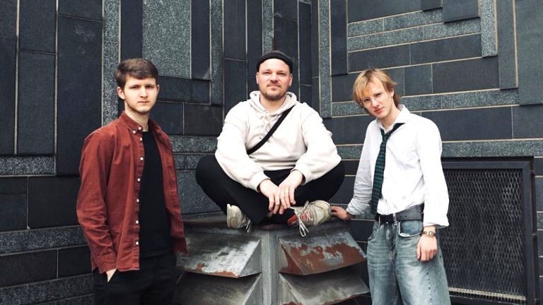 Foto des ernst blickenden Trios vor einer Wand, die mit schwarzen Steinplatten verkleidet ist