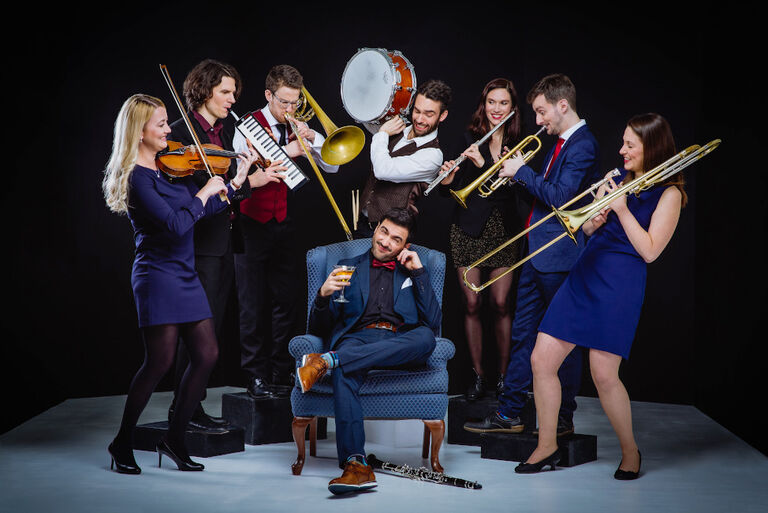 Gruppenfoto des Ensembles, das mit seinen Instrumenten um eine Person im Sessel herum steht, die sich ein Ohr zuhält.