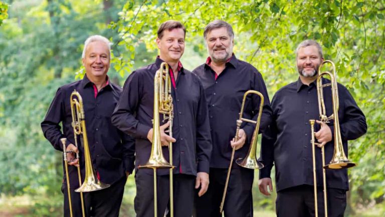 Die vier Musiker stehen mit ihren Posaunen in einem sonnigen Wald.