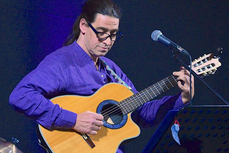 Der Musiker mit Pferdeschwanz und Hornbrille spielt vor einem Mikrofon Gitarre