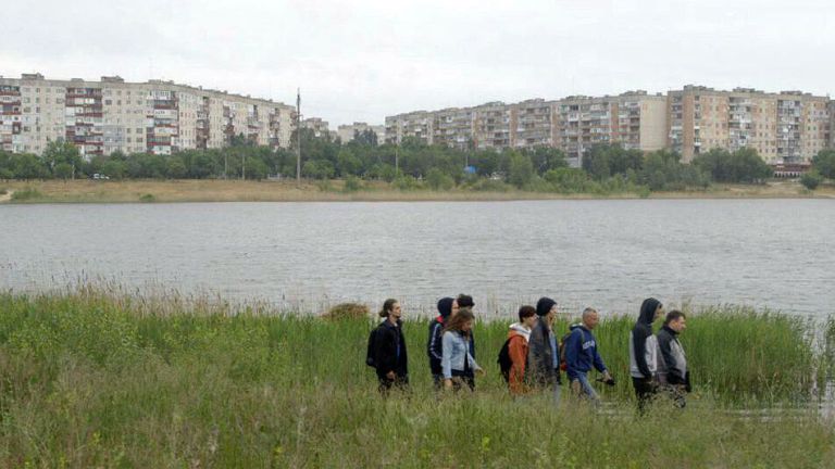 Eine Gruppe Menschen läuft durch hohes Gras an einem Fluß entlang, an dessen anderem Ufer man eine große Plattenbaussiedlung sieht.