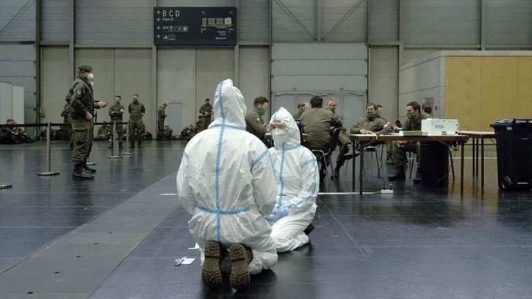 Zwei Personen in weißen Schutzanzgen und mit Masken knien in einer fast leeren Halle auf dem Boden. Im Hintergrund sieht man Uniformierte, die ebenfalls Masken tragen.