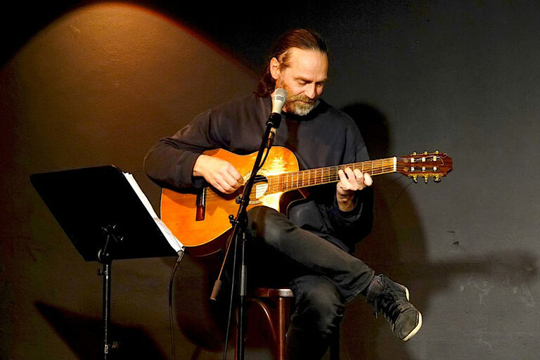 Der Liedermacher sitzt auf einem Stuhl und spielt Gitarre, vor sich ein Mikrofon und einen Notenständer.
