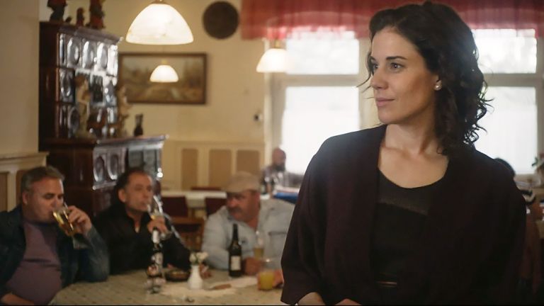 Filmszene, in der die Hauptdarstellerin im Gastraum einer dörflichen Kneipe steht, während drei Männer hinter ihr am Tisch trinken