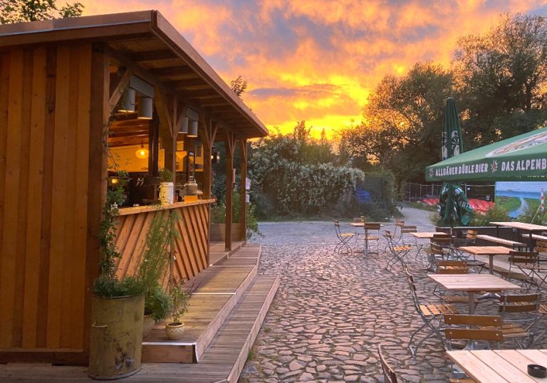 Ein Biergarten mit Holzstühlen und -tischen, einem Verkaufskiosk und dem Sonnenuntergang im Hintergrund