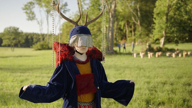 Eine Schauspielerin steht auf einer Wiese und trägt auf dem Kopf ein Geweih, an dem Perlenketten hängen.