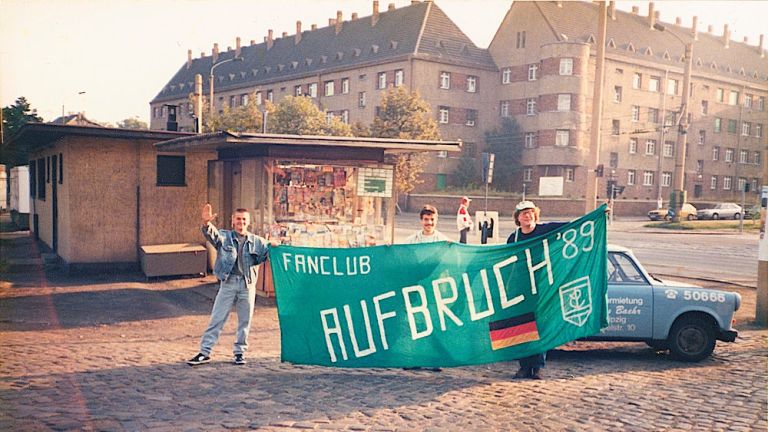 Drei Fans stehen vor einem Trabi und halten ein grünes Banner mit der Aufschrift "Fanclub Aufbruch '89" und dem Logo von Chemie Leipzig