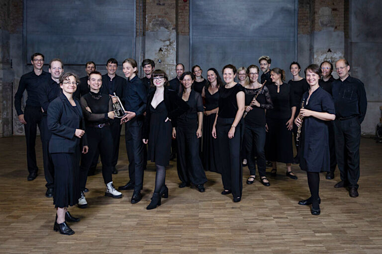Gruppenfoto der lächelnden Musikerinnen und Musiker in einem großen, etwas düsteren Saal