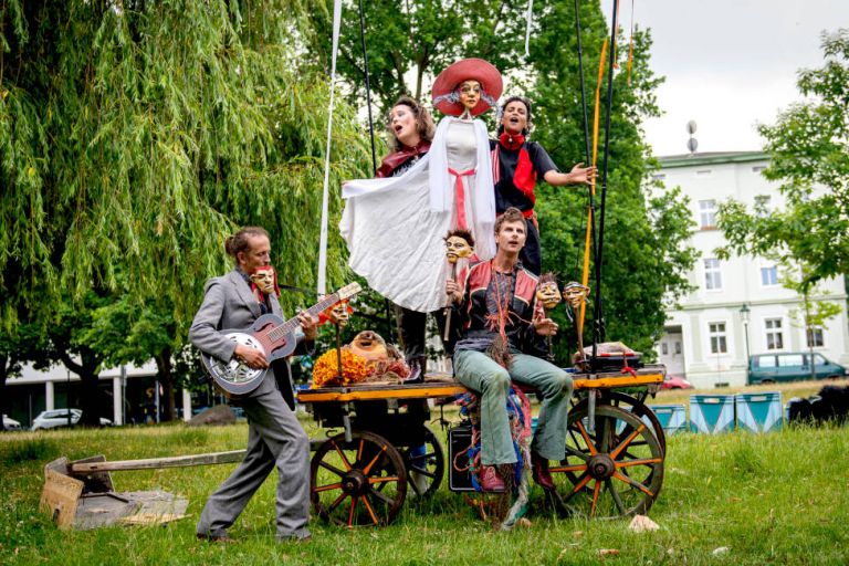 Zwei Schauspielerinnen und zwei Schauspieler sind mit Puppen und Instrumenten auf einem flachen Wagen