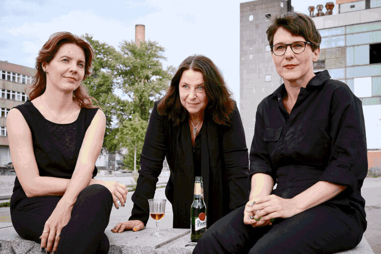 Drei Frauen in schwarzer Kleidung trinken Bier an einer Mauer.