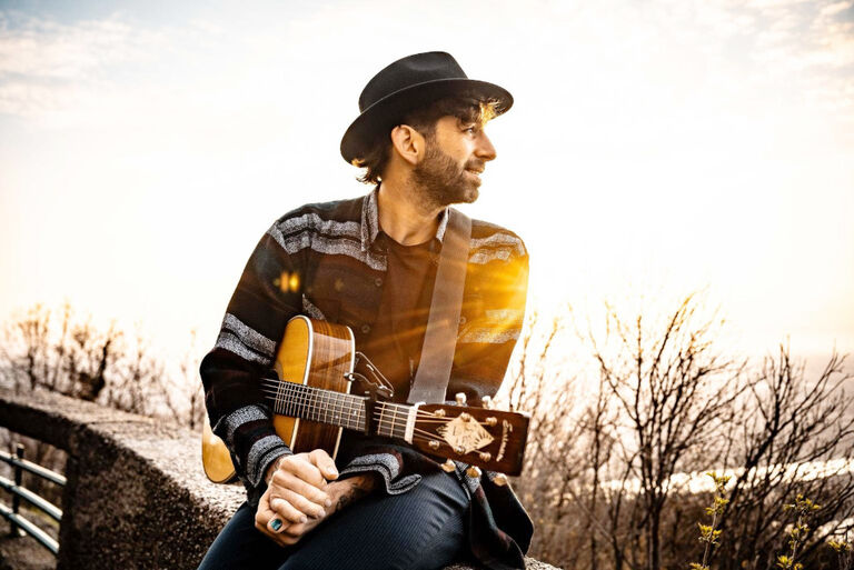 Der Musiker sitzt mit seiner Gitarre auf einer Anhöhe über einem Fluss und schaut in die Ferne.