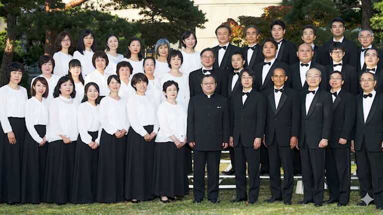 Gruppenfoto der ernst blickenden Sängerinnen und Sänger in einheitlicher Kleidung. Die Männer tragen einen schwarzen Anzug und Fliege, die Frauen eine weiße Bluse zu einem schwarzen Rock.