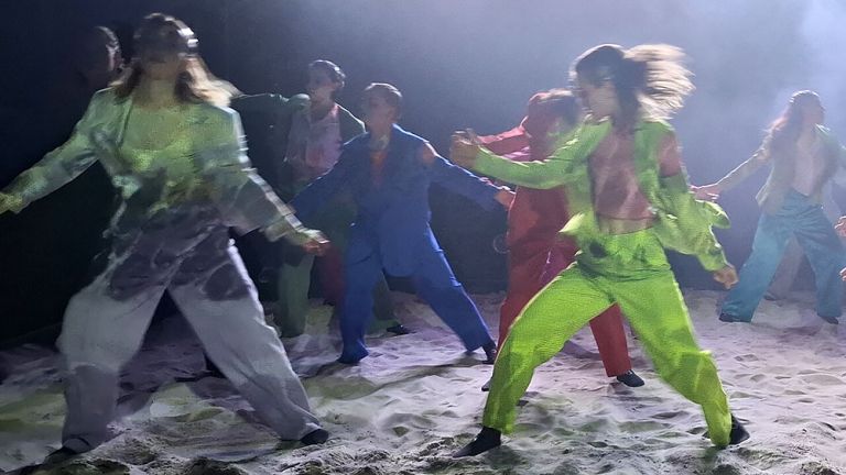 Szene mit viel Beweggung von mehreren jungen Schauspielerinnen und Schauspielern aud sandigem Boden.