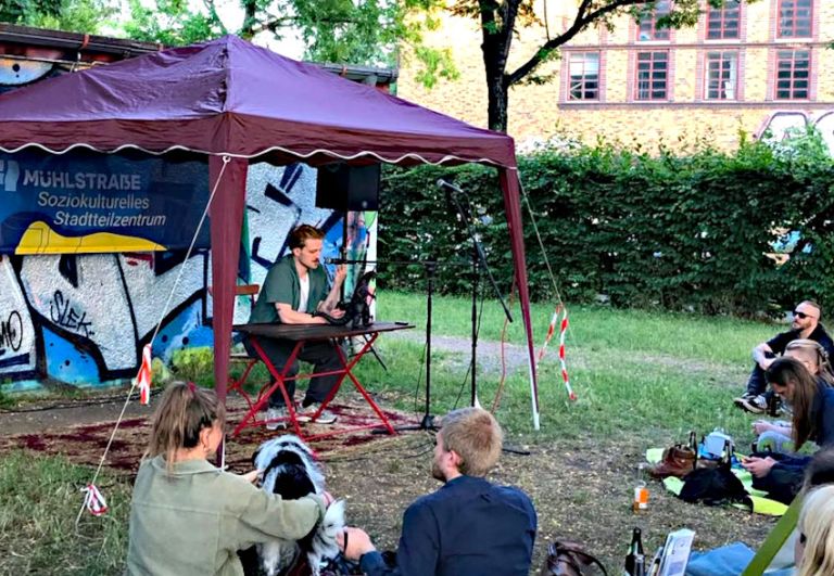 In einem offenen Pavillion sitzt ein Mann und liest etwas vor. Vor ihm auf einer Wiese sitzt das Publikum.