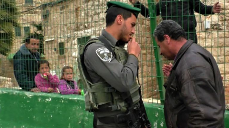 Filmszene in der sich ein Soldat und ein Mann in Zivil unterhalten, durch einen Drahtzaun sieht man einen weiteren Mann und zwei Kinder