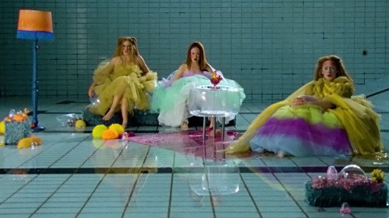 In einem leeren Schwimmbecken sitzen drei Schauspielerinnen in farbenfrohen Kleidern.