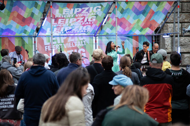 Viele Menschen stehen vor einer Bühne, auf der ein Konzert läuft. Hinter Bühne hängen Banner mit der aufschrift "Leipzig feiert Vielfalt".