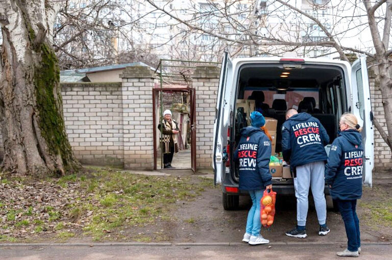 Ein Hilfs-Team holt Lebensmittel aus einem Transporter. Im Hintergrund wartet eine alte Frau vor ihrer Tür.