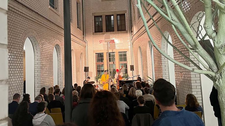 In einem Lichthof sitzen und stehen viele Menschen und verfolgen den Auftritt eines Musikensembles.