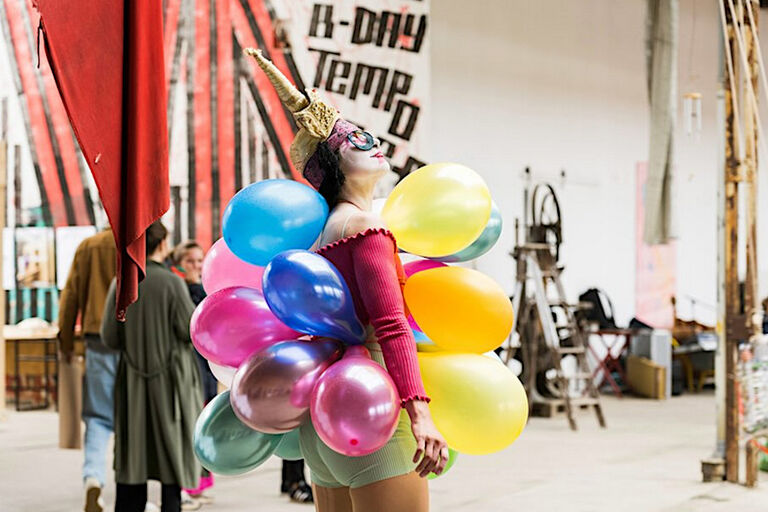 Eine Frau im Clownskostüm läuft mit vielen Loftballons durch eine Ausstellungshalle.