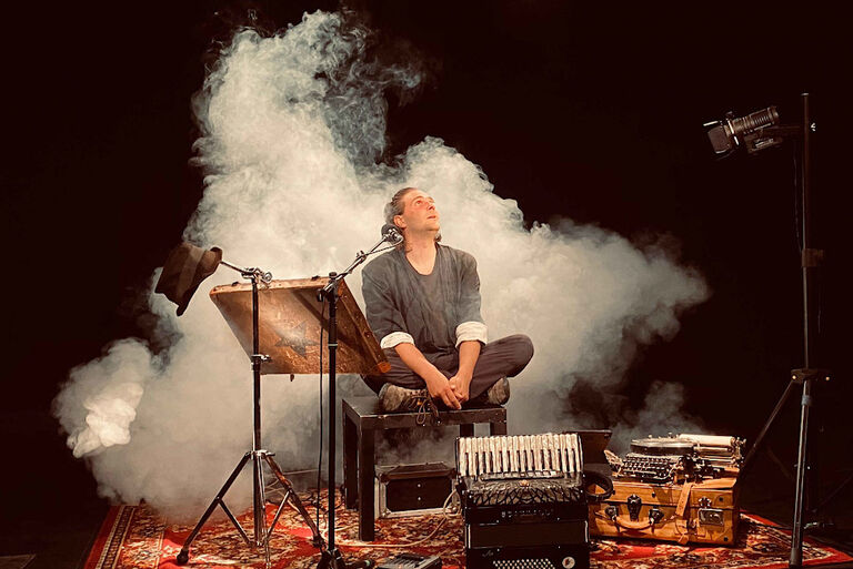 Ein Künstler sitzt im Schneidersitz auf einem Hocker. Um ihn herum stehen ein Mikrofon, ein Akkordean, ein Koffer mit einem Stapel Heften und hinter ihm ist eine Nebelwolke zu sehen.