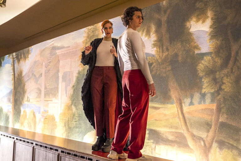 Eine junge Frau und ein junger Mann stehen in roten Hosen und weißem T-Shirt vor einem großen Wandbild mit einem Waldmotiv