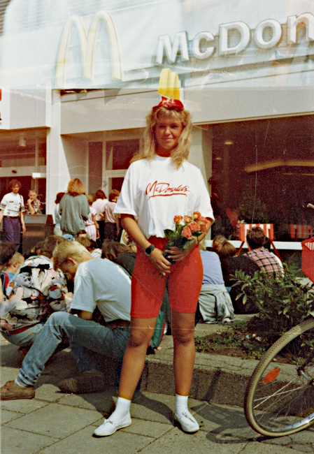 Eine junge Frau steht in kurzen roten Leggins und einem Mc Donalds T-Shirt vor einer Mc Donalds Filliale.