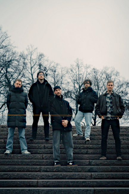 Gruppenfoto der Band auf einer Treppe vor einer herbstlichen Kuliss aus kahlen Bäumen