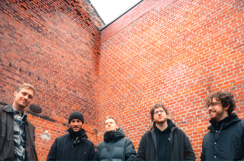 Gruppenfoto der Band in Wintersachen vor einer unverputzten Ziegelwand