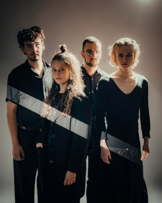 Gruppenfoto der schwarz gekleideten Band im Halbdunkel, in das eine Lichtstreifen fällt