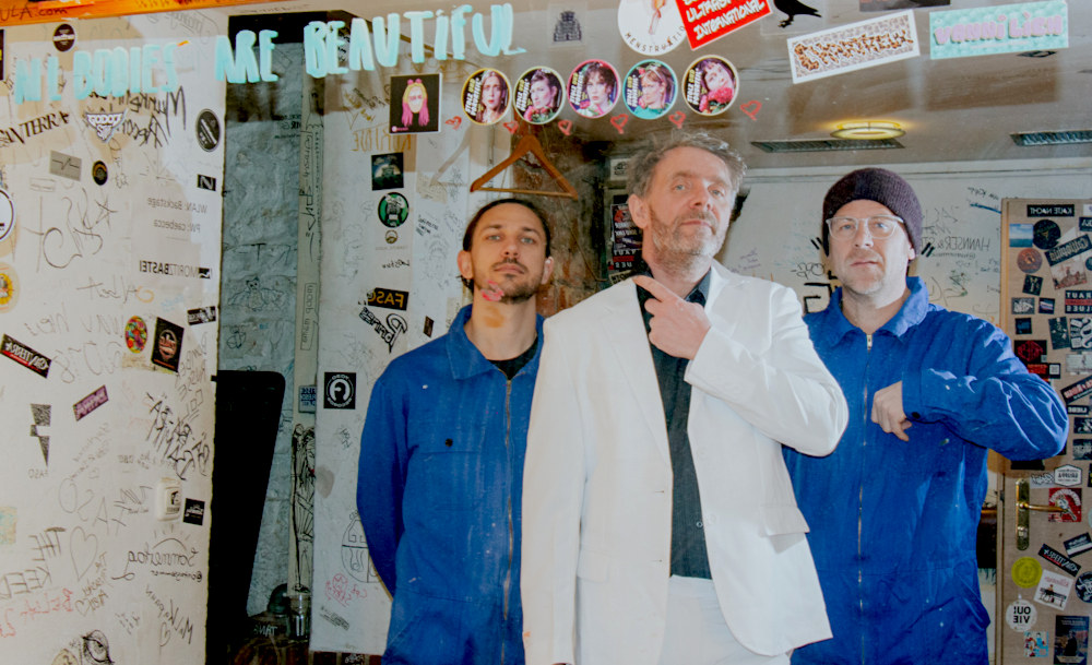 Gruppenfoto, auf dem der Sänger einen weißen Anzug trägt und die beiden anderen Musiker einen blauen Overall