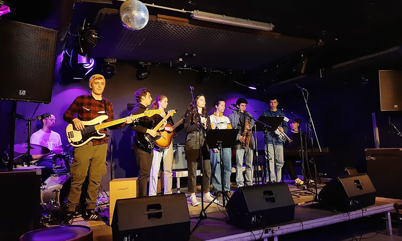 Acht Jugendliche stehen bei einem Konzert mit verscheidenen Instrumenten auf der Bühne.