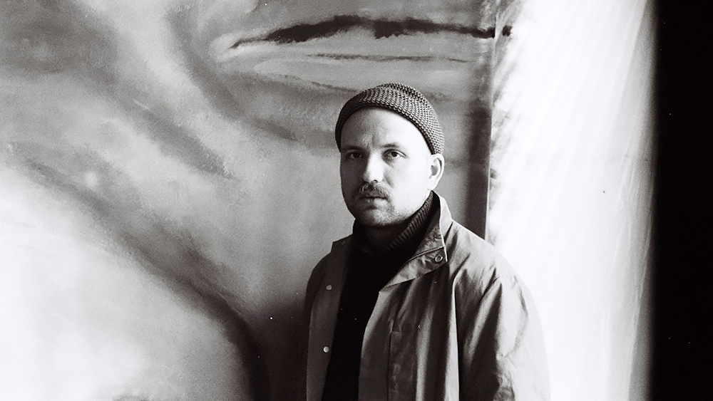 Der Künstler steht mit Jacke und Strickmütze vor einem großen Gemälde.