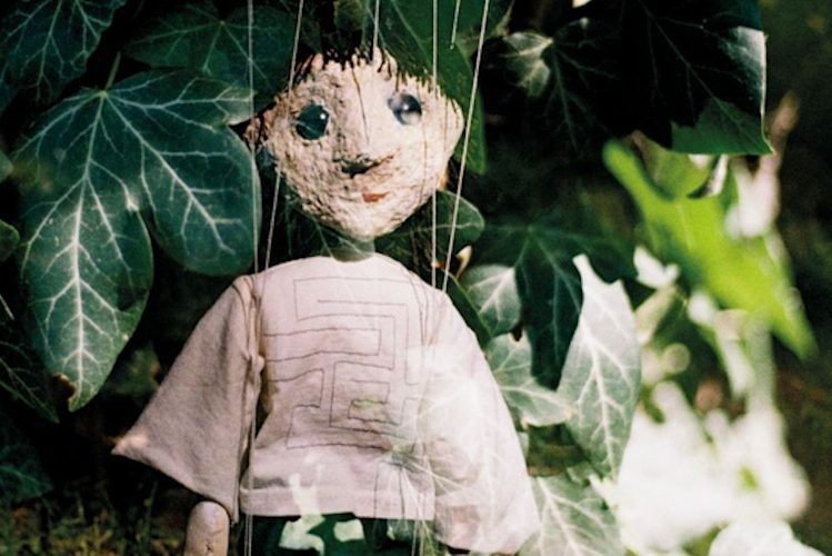 Eine Marionette mit hellem Gesicht und Glasaugen vor den Blättern einer Grünpflanze