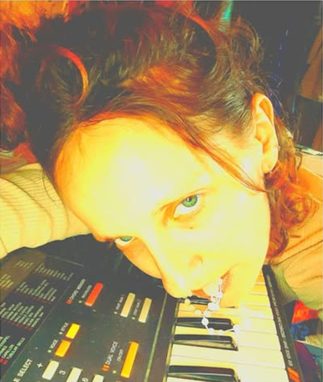 Foto der Künstlerin, die mit ihrem Kopf und einer Perlenkette im Mund auf der Tastatur eines Keyboards liegt