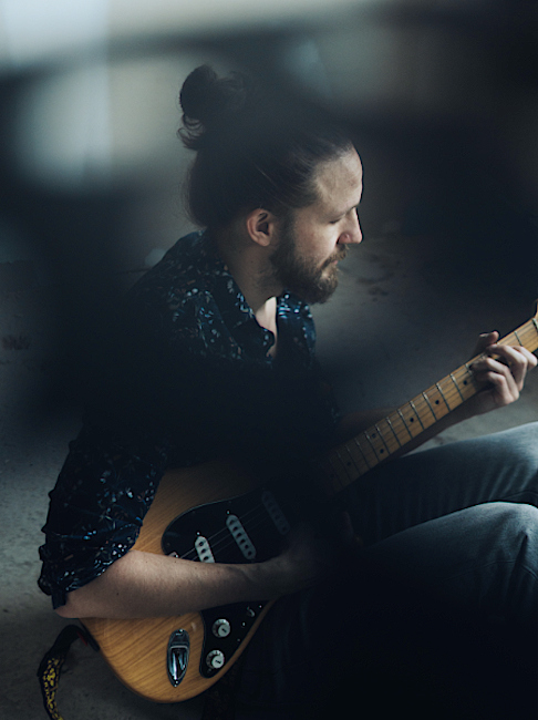 Der Musiker sitzt auf dem Boden und spielt mit konzentriertrm Gesichtausdruck E-Gitarre.