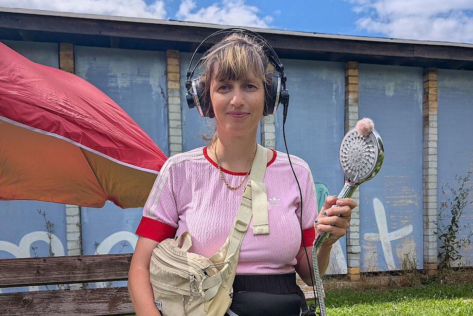 Eine junge Frau mit Kopfhörern sitzt auf einer Bank vor einem roten Sonnenschirm und hält einen Duschkopf wie ein Mikrofon in der Hand.