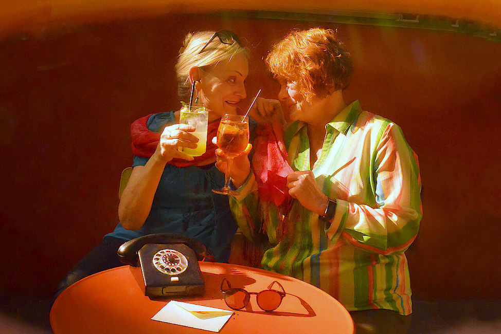Zwei ältere Frauen sehen sich liebevoll an und prosten sich mit Cocktails zu.