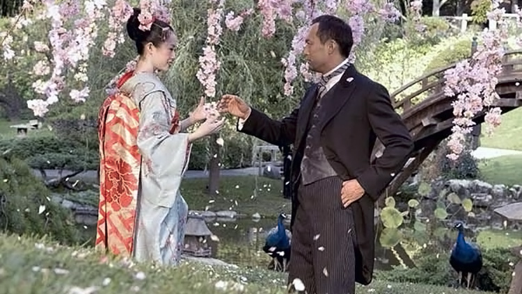 Eine junge Geisha steht in einem Garten voller Kirschblten und mit Pfauen einem Mann in feinem Anzug gegenüber.