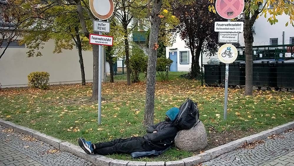 Ein Mann schläft, seinen Kopf an seinen Rucksack gelehnt auf einer kleinen Wiese mit Verkehrszeichen.