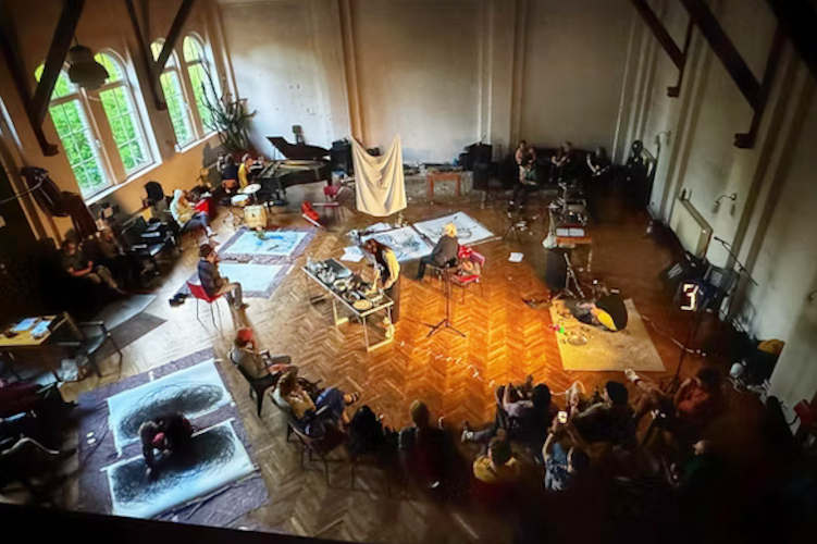 In einem großen Raum sind verschiedene Künstler am Werk, wie Maler oder Musiker, es wird gekocht und viele Menschen schauen sich die unterschiedlichen Darbietungen an.