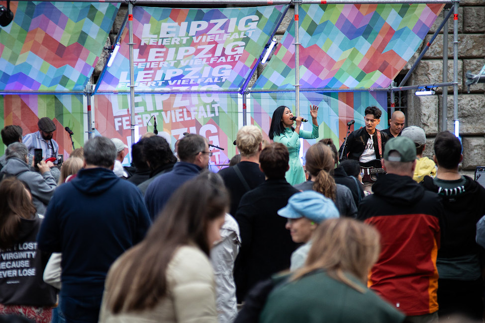Viele Menschen stehen vor einer Bühne, auf der ein Konzert läuft. Hinter Bühne hängen Banner mit der aufschrift "Leipzig feiert Vielfalt".